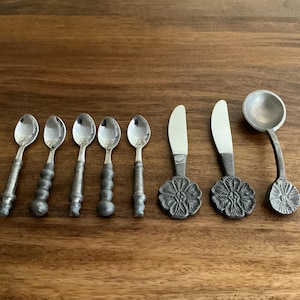 May include: A collection of silver-toned serving utensils, including five small spoons with decorative handles, two butter knives with floral bases, and a small ladle. The utensils are arranged on a wooden surface.