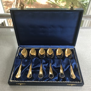 May include: A set of six gold-plated spoons in a blue velvet-lined box. The spoons are arranged in a row and have a classic design.
