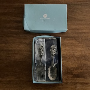 May include: A boxed set of silver-toned serving utensils, including a fork and spoon, with ornate floral and leaf detailing on the handles. The box is light blue and has the text "JHB SILVER COMPANY".