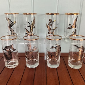May include: Set of eight clear glass tumblers with gold rims. Each tumbler features a different bird illustration, including ducks, geese, and a pheasant. The tumblers are arranged in two rows on a wooden surface.