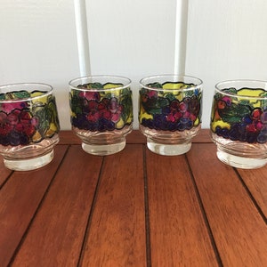 May include: Four clear glass tumblers with a grape design. The glasses have a yellow background with green leaves, purple grapes, and blue accents.