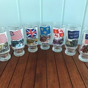 May include: Set of six clear glass drinking glasses with colorful flags and historical text printed on them. The flags include the American flag, the British flag, the French flag, the flag of the Kingdom of Jerusalem, the flag of the United States Navy, and the flag of the Grand Union.