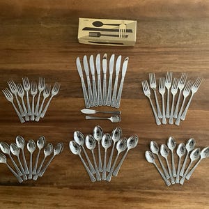 May include: A collection of vintage silverware, including forks, knives, spoons, and butter knives, arranged on a wooden surface. The silverware has a decorative pattern and is displayed in multiple sets. A box with silverware illustrations is also present.