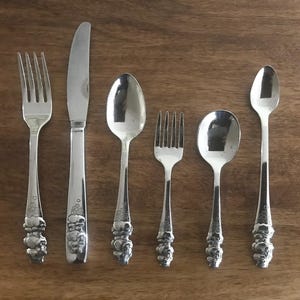 May include: A set of six silver-colored flatware pieces arranged on a wooden surface. The set includes two forks, two spoons, and a knife. The handles have a decorative floral design.