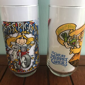 May include: Two vintage glass drinking glasses with colorful Muppet characters. The first glass features Miss Piggy riding a motorcycle with the text "Miss Piggy" above her. The second glass features Kermit the Frog and Fozzie Bear with the text "Kermit the Frog" and "The Great Muppet Caper!"