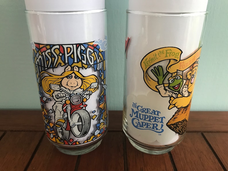 May include: Two vintage glass drinking glasses with colorful Muppet characters. The first glass features Miss Piggy riding a motorcycle with the text "Miss Piggy" above her. The second glass features Kermit the Frog and Fozzie Bear with the text "Kermit the Frog" and "The Great Muppet Caper!"