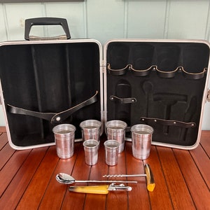 May include: Open vintage cocktail set with a silver-colored case, black interior, and a handle. The case contains several silver cups, a spoon, a corkscrew, and other bar tools. The set is displayed on a wooden surface.
