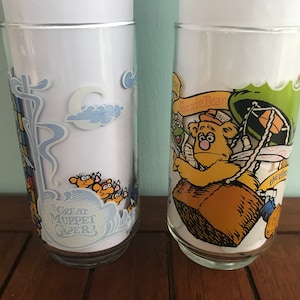 May include: Two vintage glass drinking glasses with cartoon illustrations. The first glass features a blue and white design with the text "The Great Muppet Caper!" The second glass features a yellow bear with a green hat and the text "Fozzie Bear".