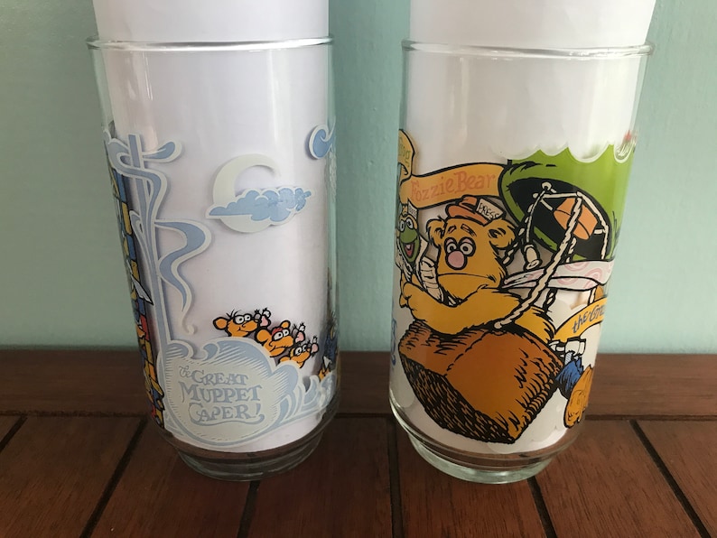 May include: Two vintage glass drinking glasses with cartoon illustrations. The first glass features a blue and white design with the text "The Great Muppet Caper!" The second glass features a yellow bear with a green hat and the text "Fozzie Bear".