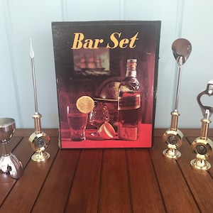 May include: A vintage bar set in its original box. The set includes a jigger, a bottle opener, a cocktail stirrer, and a cocktail pick. The box has a picture of a bottle of liquor, a globe, and a lemon slice.