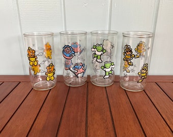 May include: Set of five clear glass drinking glasses with winter-themed cartoon characters. The glasses feature a bear, a frog, and a penguin in various winter activities, such as ice skating and sledding. The glasses have a snowflake pattern.
