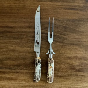 May include: A set of two vintage carving utensils with stag horn handles. The knife has a silver blade with an etched design of a deer and other wildlife. The fork has a silver tines and a stag horn handle.