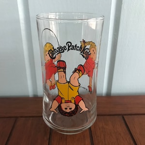 May include: A clear glass with a Cabbage Patch Kids design. The design features two children, one boy and one girl, playing jump rope. The text "Cabbage Patch Kids" is printed in black on a white background.