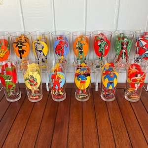 May include: Set of 14 vintage glass tumblers featuring various DC Comics superheroes, including Superman, Batman, Robin, Wonder Woman, and The Flash. Each tumbler has a different character and a yellow background.