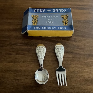 May include: A vintage baby spoon and fork set with a box. The spoon and fork are silver-colored, each featuring a dog design at the handle's top. The box is blue and yellow, with the text "ANDY and SANDY BABY SPOON and FORK THE SHAGGY PALS".