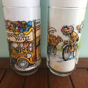 May include: Two vintage glass tumblers with Sesame Street characters. The first tumbler features Big Bird driving a yellow bus that says "Happiness Hotel". The second tumbler features Cookie Monster riding a bicycle.