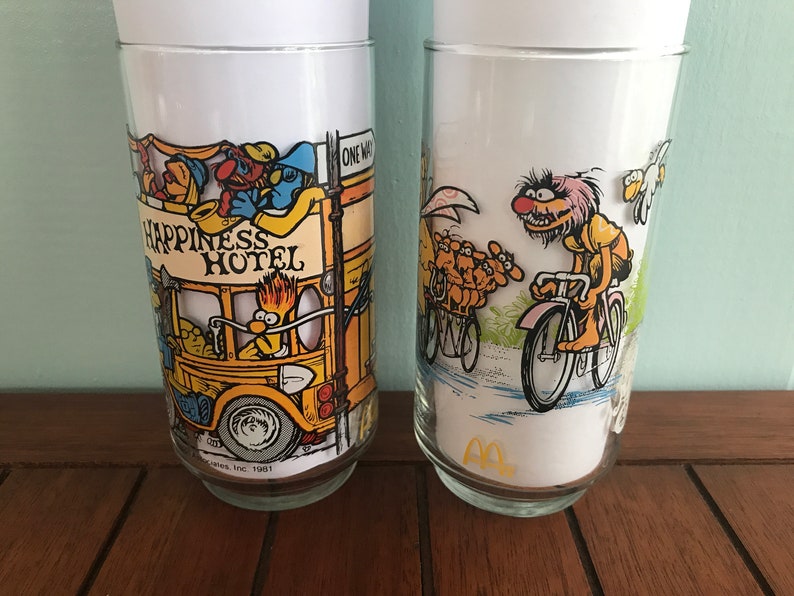 May include: Two vintage glass tumblers with Sesame Street characters. The first tumbler features Big Bird driving a yellow bus that says "Happiness Hotel". The second tumbler features Cookie Monster riding a bicycle.