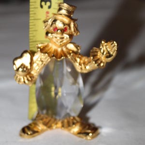 May include: A gold-colored clown figurine with red eyes and a clear, faceted body. The clown is wearing a hat and a ruffled collar. The figurine is approximately 3 inches tall.