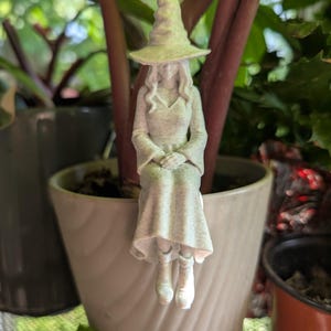 May include: A light-colored, speckled figurine of a seated witch wearing a pointed hat, a long-sleeved top, and a skirt. The figure has long hair and is depicted in a sitting position with hands resting in the lap, designed to hang from a pot edge.