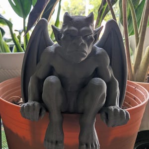 May include: A dark gray, 3D-printed gargoyle figurine with pointed ears, horns, and folded wings. The creature is depicted in a seated, crouching position with its hands and feet resting over the edge of a large, orange plastic plant pot.