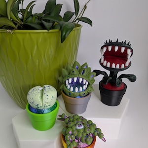May include: A collection of four small potted plant figurines. One is a pale green split succulent, one is a spiky green monster with white teeth, one is a black monster with a wide open mouth and white teeth, and one is a green multi-eyed creature.