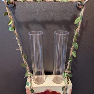 May include: A decorative wall hanging with two clear glass tubes. The tubes are suspended from a twine and faux leaf garland attached to silver hooks. The base is a rectangular block with red and cream-colored paint.