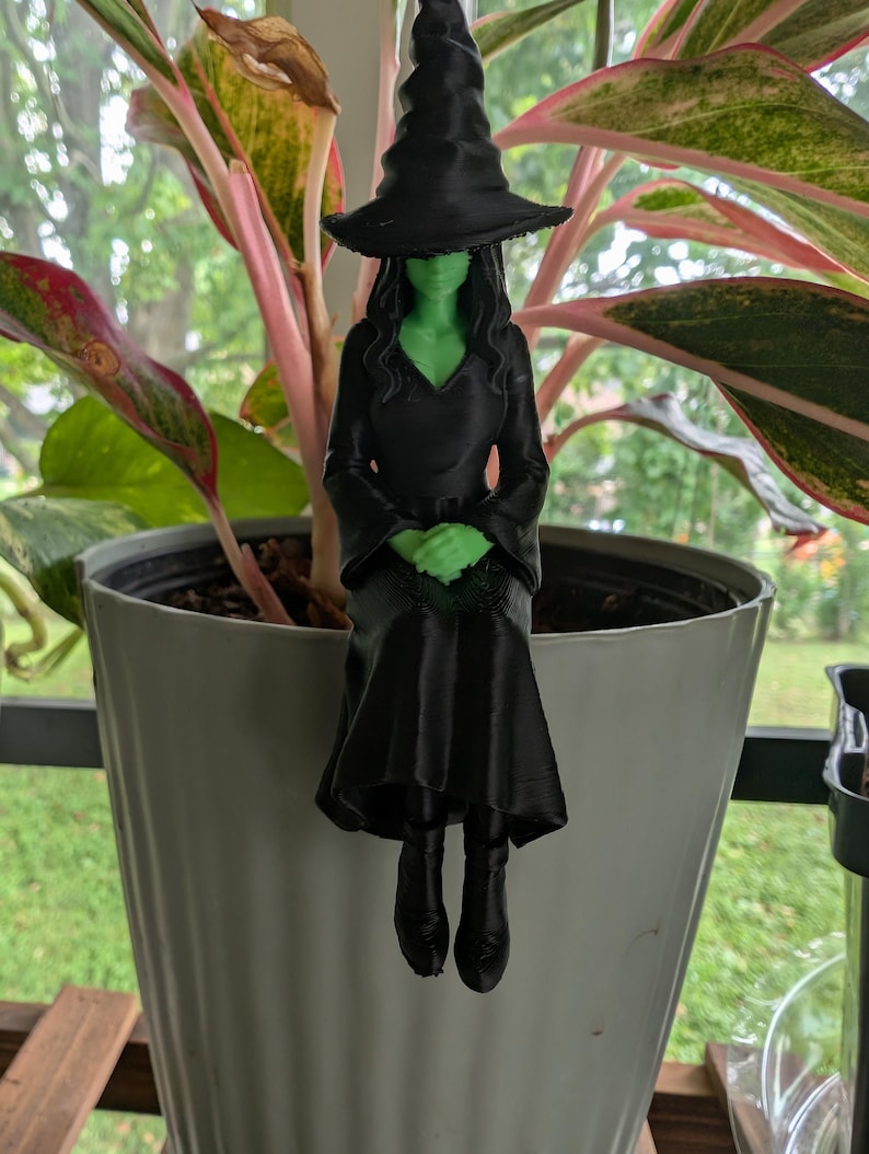 May include: A 3D printed witch figurine with green skin and black hair, wearing a black pointed hat, a long-sleeved black dress, and black boots. The figure is designed to sit on the edge of a container, with hands folded in the lap.