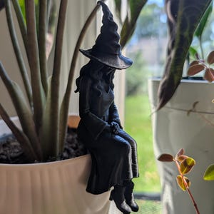 May include: A matte black figurine of a witch wearing a pointed hat and a long dress with flared sleeves. The figure is depicted in a seated position with legs dangling, designed to perch on the edge of a container or a plant pot rim.