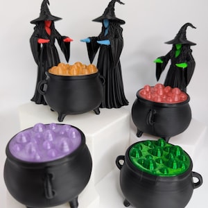 May include: Three black witch figurines wearing pointed hats and long robes, each with distinct red, blue, or green skin and hands. Four black cauldrons are filled with bubbling liquid in orange, red, purple, and green colors respectively.