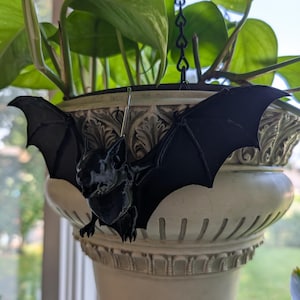 May include: A black bat-shaped ornament with outstretched wings, suspended by a silver-colored metal hook. The bat is attached to the rim of a cream-colored, fluted ceramic planter that contains a green leafy plant with heart-shaped foliage.