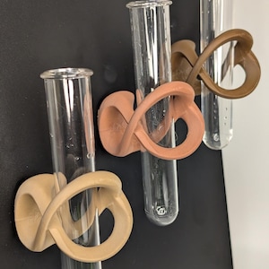 May include: Three clear glass test tubes with decorative holders. The holders are in shades of tan and brown, featuring an abstract, looped design. The test tubes are mounted on a black surface, with a white background visible on the right.
