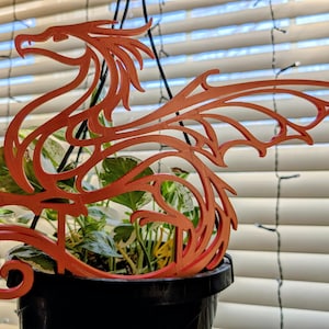 May include: A decorative, orange dragon silhouette, likely 3D-printed, is suspended above a potted plant. The dragon's design features intricate cutouts and a stylized form. The plant is in a black pot, and the background includes blinds and string lights.