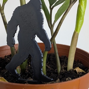 May include: A black, flat, silhouette-style figure of a Bigfoot creature standing upright. The figure is positioned within a brown plastic plant pot containing a green plant with multiple stems and leaves, partially obscured by the plant stalks.