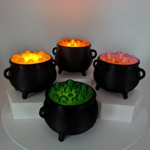 May include: Four black three-legged cauldrons with side handles. Each cauldron contains a translucent, textured substance on top. Two cauldrons glow with a bright yellow-orange light, one glows with a soft pink light, and one glows with a vibrant green light.