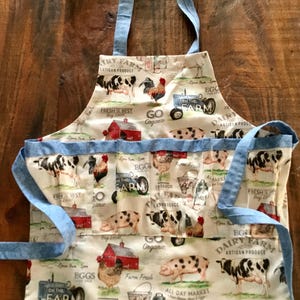 May include: A white apron with a farm-themed pattern featuring cows, chickens, pigs, and red barns. The apron has light blue trim and ties. Text includes "Dairy Farm Artisan Produce" and "Fresh is Best Buy Local".