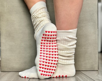 Puede incluir: Un par de calcetines blancos con agarres en forma de corazón rojos en las suelas. Los calcetines están doblados en los tobillos, revelando los agarres.