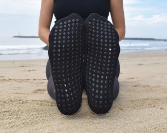 May include: A pair of black grippy socks with a textured pattern on the bottom. The socks are worn by a person sitting on a sandy beach.