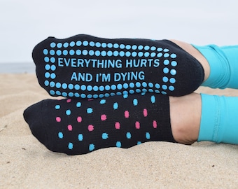 May include: Black socks with blue and pink polka dots and the text "EVERYTHING HURTS AND I'M DYING" on the soles. The socks are worn on a sandy beach, with blue leggings visible.