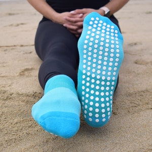 May include: A pair of blue socks with white grippy dots on the bottom. The socks are worn by a person sitting on the sand.