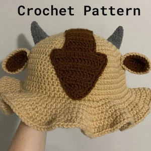 以下が含まれることがあります： ベージュ、茶色、グレーの、角と耳のような形が特徴の、かぎ針編みの帽子。つばが広く、前面に茶色の矢印型のデザインがあります。「Crochet Pattern」の文字が上部にあります。