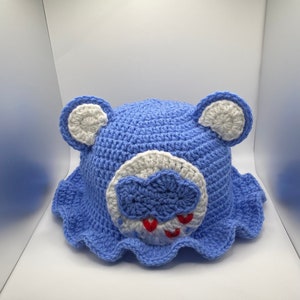 May include: A handmade blue crochet hat with white accents, featuring bear ears and a cloud design with red hearts. The hat has a scalloped brim and is designed for a child.