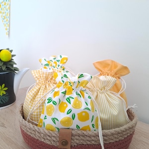 May include: A collection of drawstring bags in a woven basket. The bags feature lemon and gingham patterns in yellow and white. A decorative teapot with lemons and a banner with lemon slices are also visible.