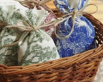 May include: A woven wicker basket holds several small fabric pouches tied with twine. The pouches feature various patterns, including a tree design in green, a floral pattern in burgundy, and a blue and white toile print.