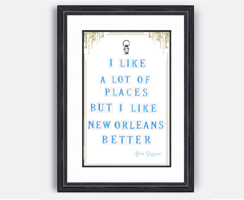 May include: A framed print with a white background and blue text that reads "I like a lot of places but I like New Orleans better - Bob Dylan". The text is in a blocky font and is surrounded by a gold frame.