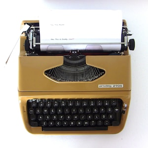 May include: A gold typewriter with a sheet of white paper in it. The typewriter has a black keyboard with letters and numbers. The typewriter says "privileg 270S" on the front.