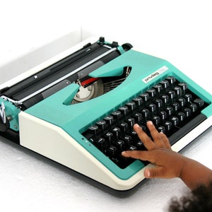 Peut inclure: Une machine à écrire turquoise vintage avec des touches noires et une base blanche. Le mot "privileg" est visible sur la machine à écrire. Une main d'enfant se dirige vers les touches. La machine à écrire est sur une surface blanche.