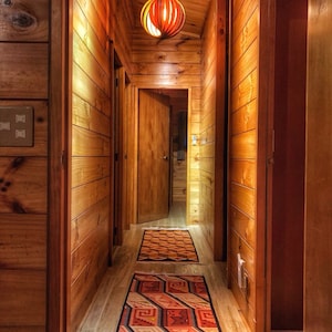 Puede incluir: Un pasillo largo y estrecho con paredes y techo de madera. El pasillo está iluminado por una lámpara colgante con una pantalla redonda de color naranja. Hay dos puertas a cada lado del pasillo, y una alfombra estampada recorre el centro.