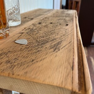May include: Close-up of a rustic wooden table top with a worn, natural finish. The table has a slightly rounded edge and a guitar pick is resting on the surface.