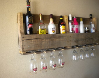 May include: A rustic wooden wall-mounted wine rack with a shelf and glass holders. The rack is filled with various bottles of liquor, including tequila, rum, vodka, and liqueur.