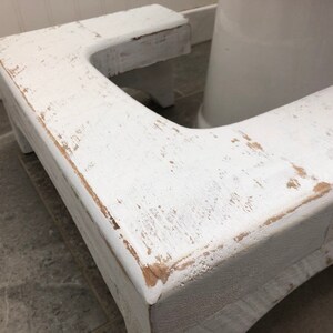 May include: White wooden toilet seat riser with a distressed finish. The riser is designed to help people reach the toilet seat more easily.